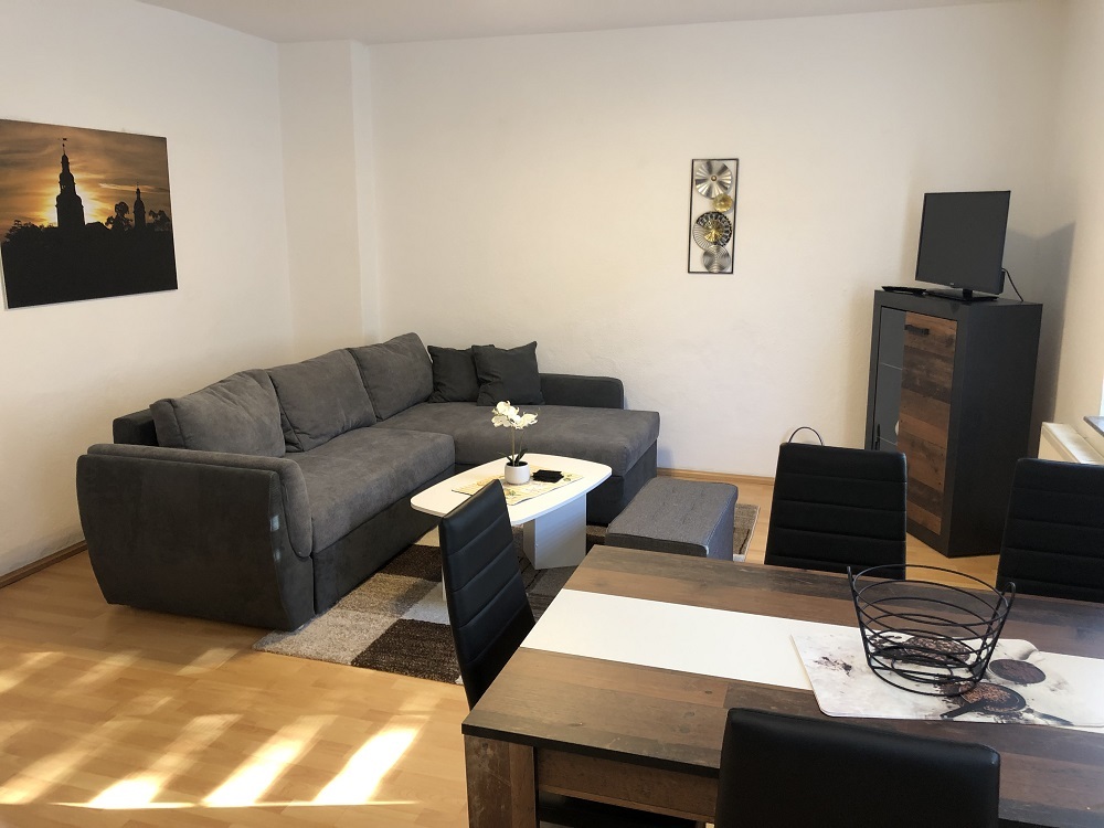 Wohnzimmer der Ferienwohnung 1 mit grauem Ecksofa, Esstisch und einem Wandbild der Sondershäuser Stadtsilhouette