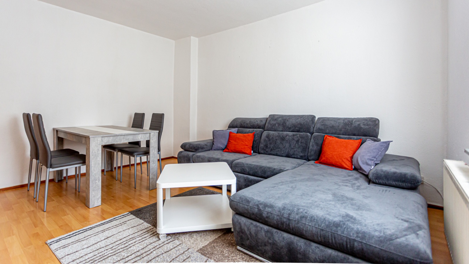 Wohnzimmer der Ferienwohnung 2 mit großem grauem Ecksofa, orangefarbenen Kissen, Couchtisch und Essplatz